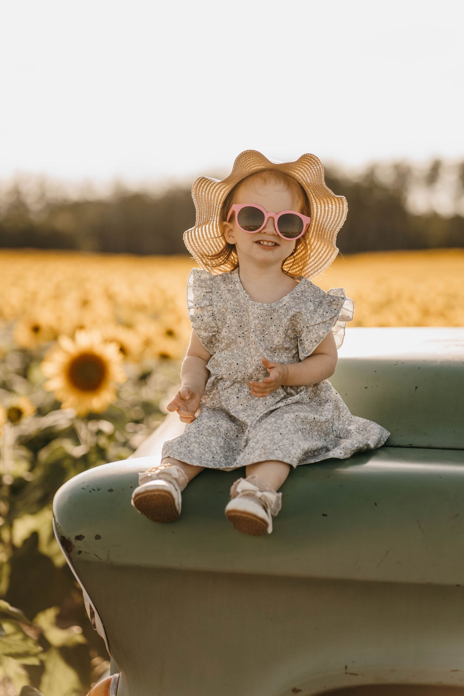 Baby wearing sunhat and floral dress sits on green surface near sunflower field at sunset.