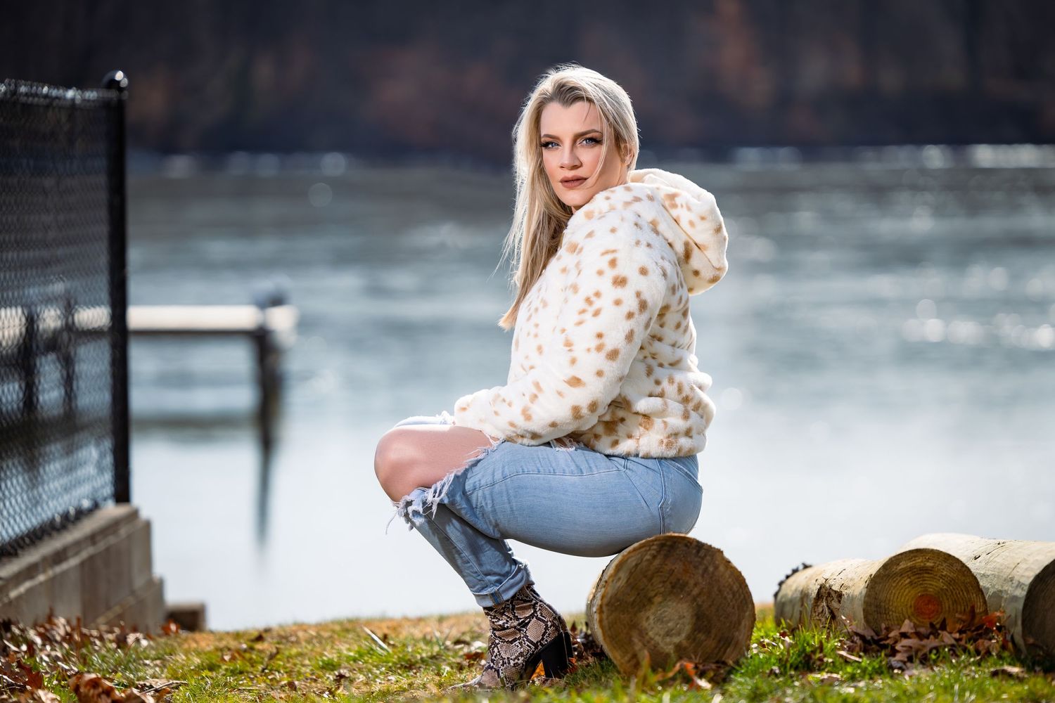 Woman in spotted hoodie and jeans sits on log by lake.