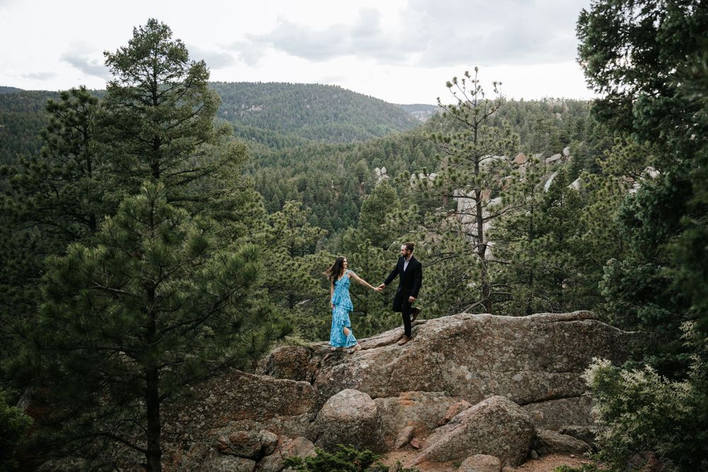 mountains boulders view man in dark suit bun woman in turquoise dress walking on boulders