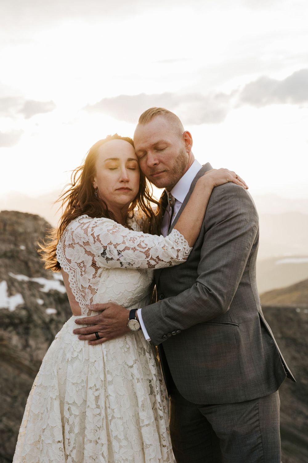 bride in boho lace dress groom in gray suit mohawk holding each other rocky mountaintop epic view colorado snow sunset
