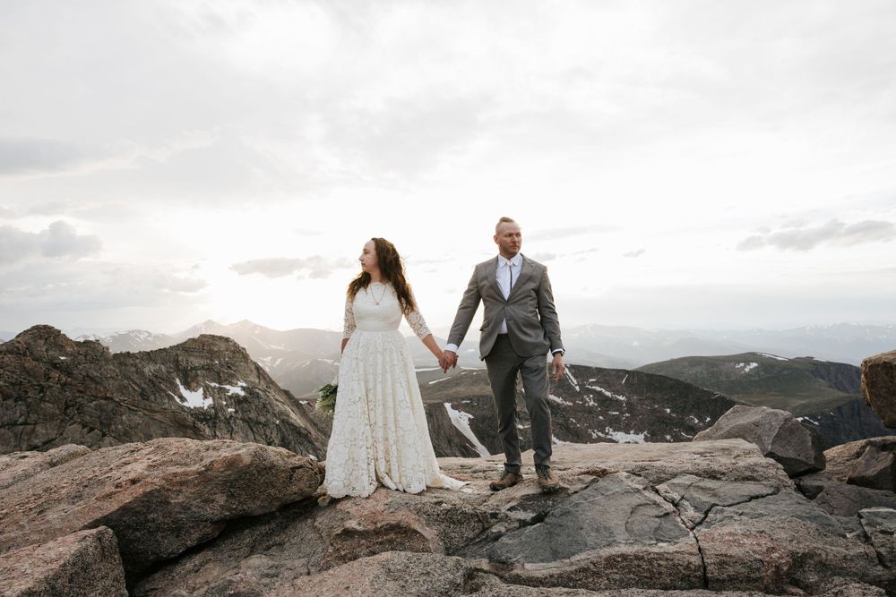bride in boho lace dress groom in gray suit mohawk holding hands standing on mountaintop epic view colorado snow sunset