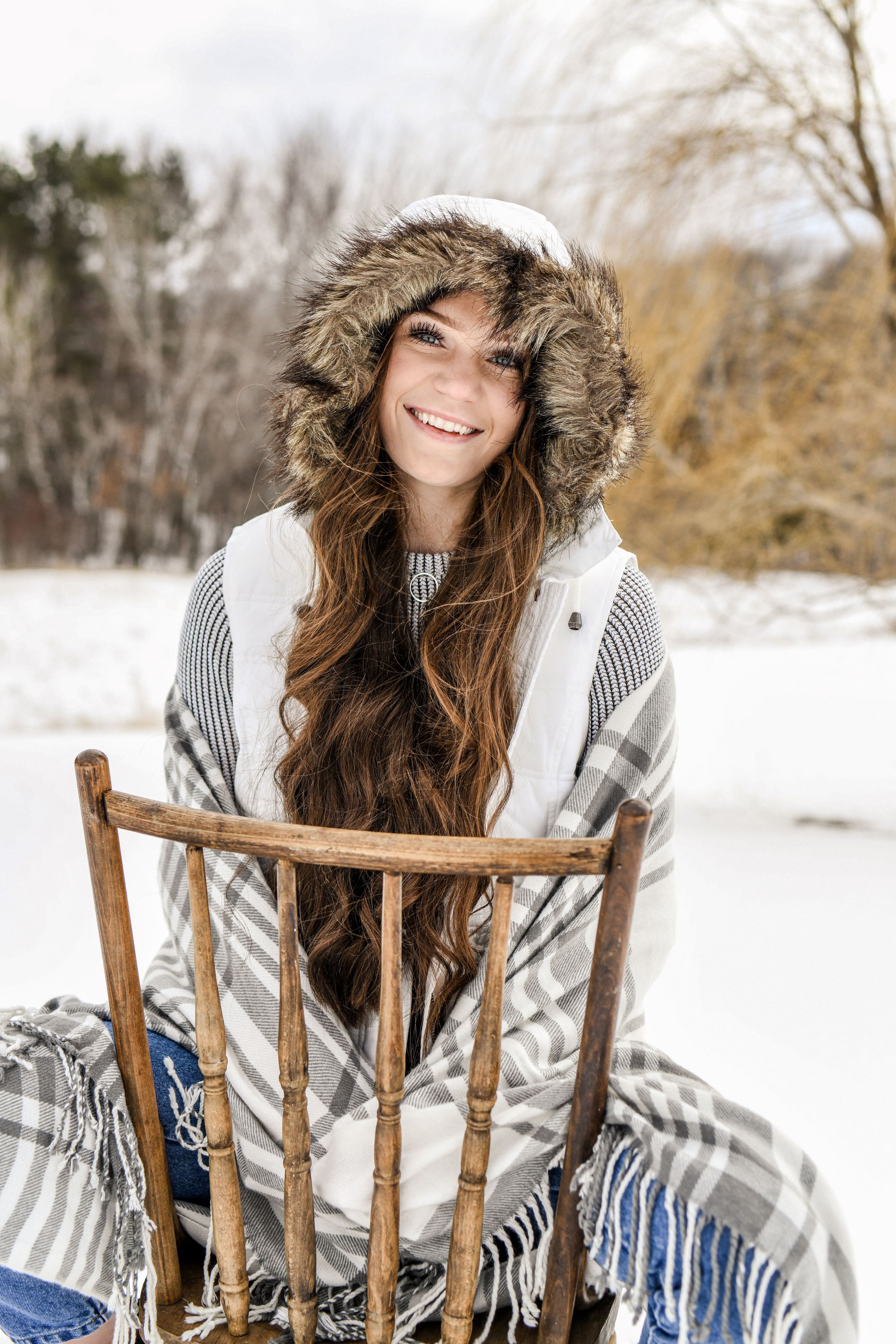 A high school senior winter portrait featuring a cozy fur-trimmed hood and sweater against a snowy outdoor backdrop with a wooden chair.
