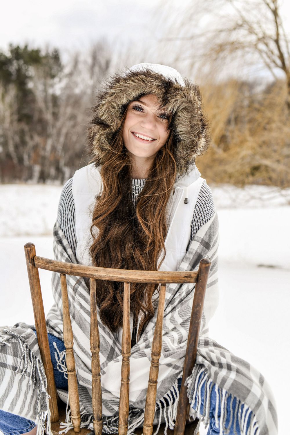 A high school senior winter portrait featuring a cozy fur-trimmed hood and sweater against a snowy outdoor backdrop with a wooden chair.
