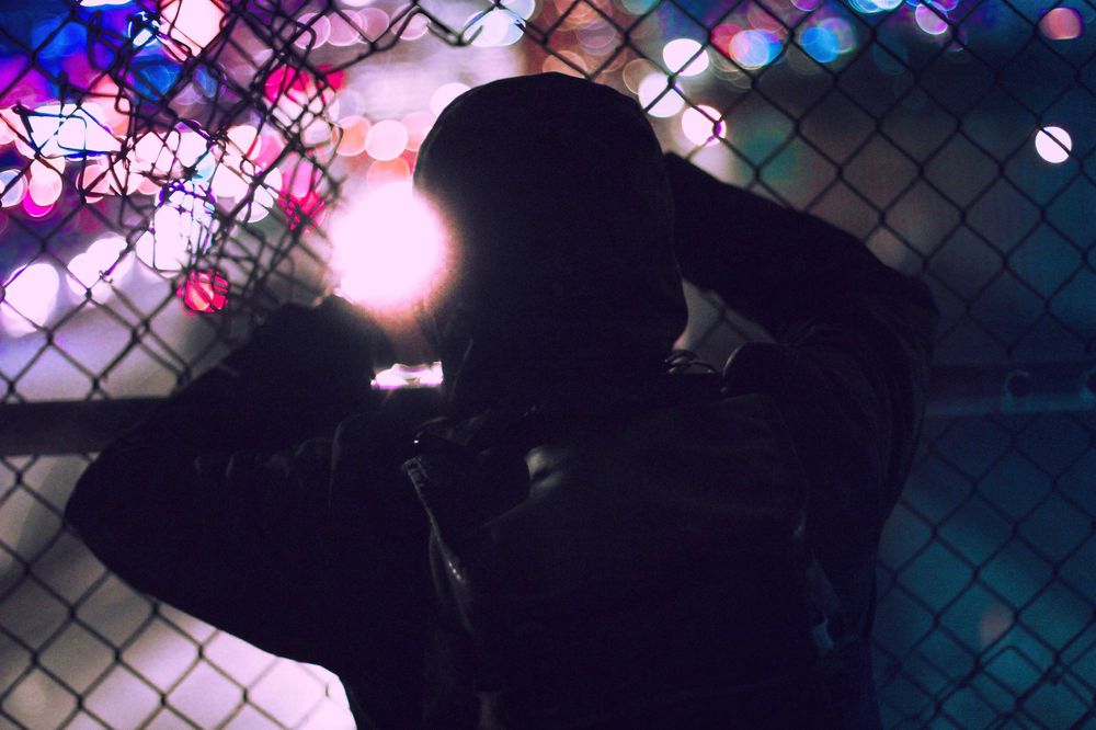 Silhouetted figure against chain-link fence with colorful bokeh lights in background.