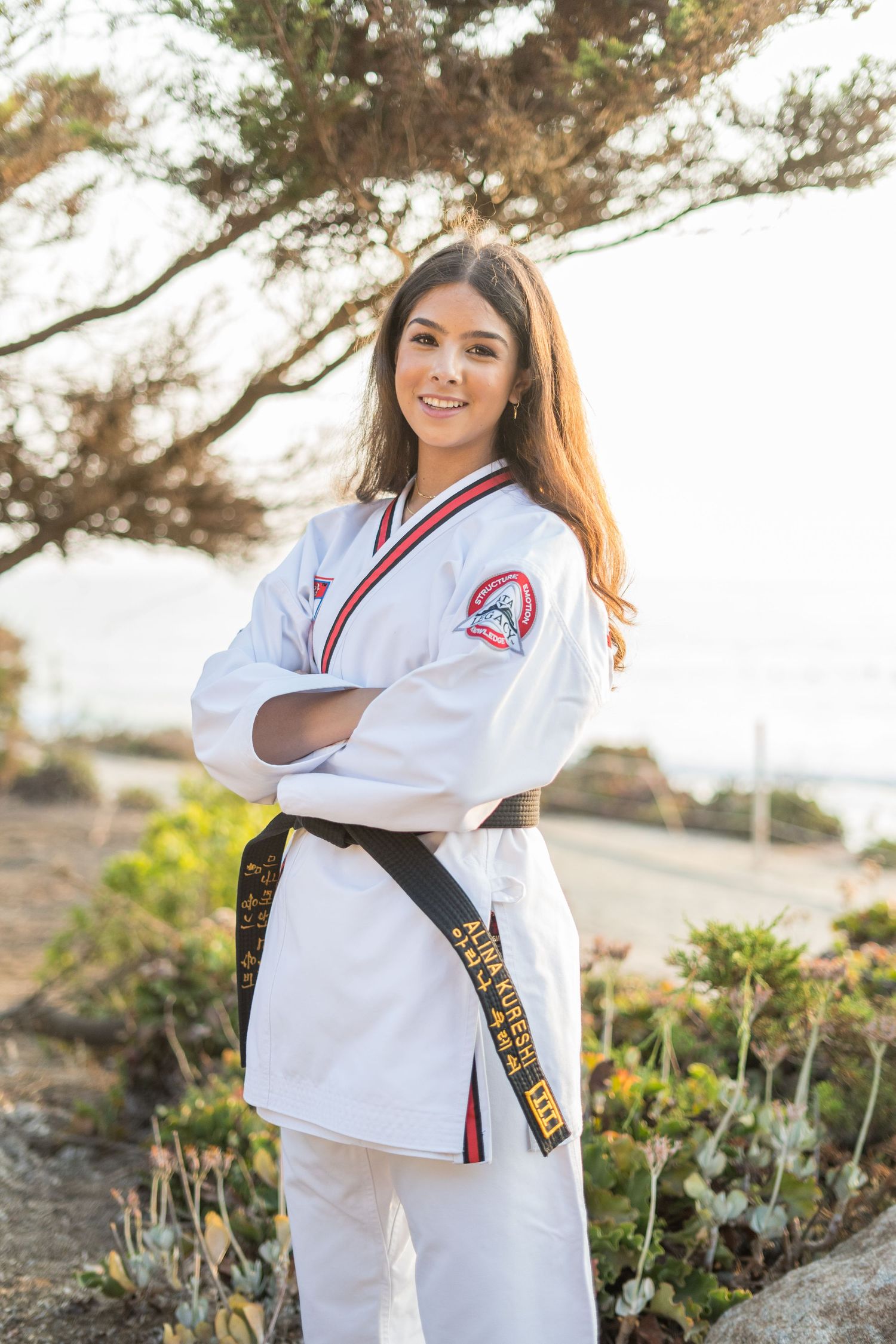 Martial artist posing outdoors in white taekwondo uniform with black belt against natural backdrop.