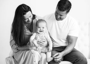one year old girl plays with her feet and smiles on mom and dad's lap