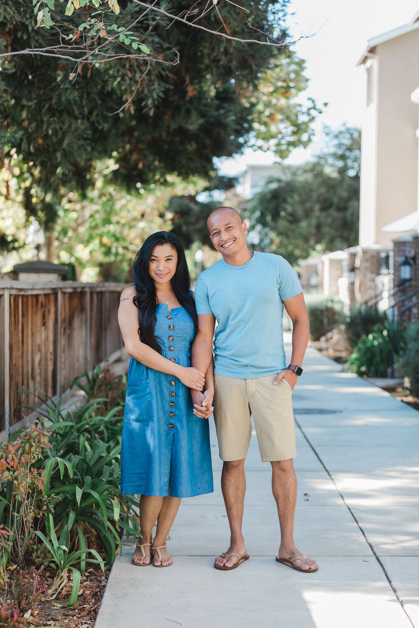Couple stands together on a sunny sidewalk wearing casual summer outfits of blue and khaki.