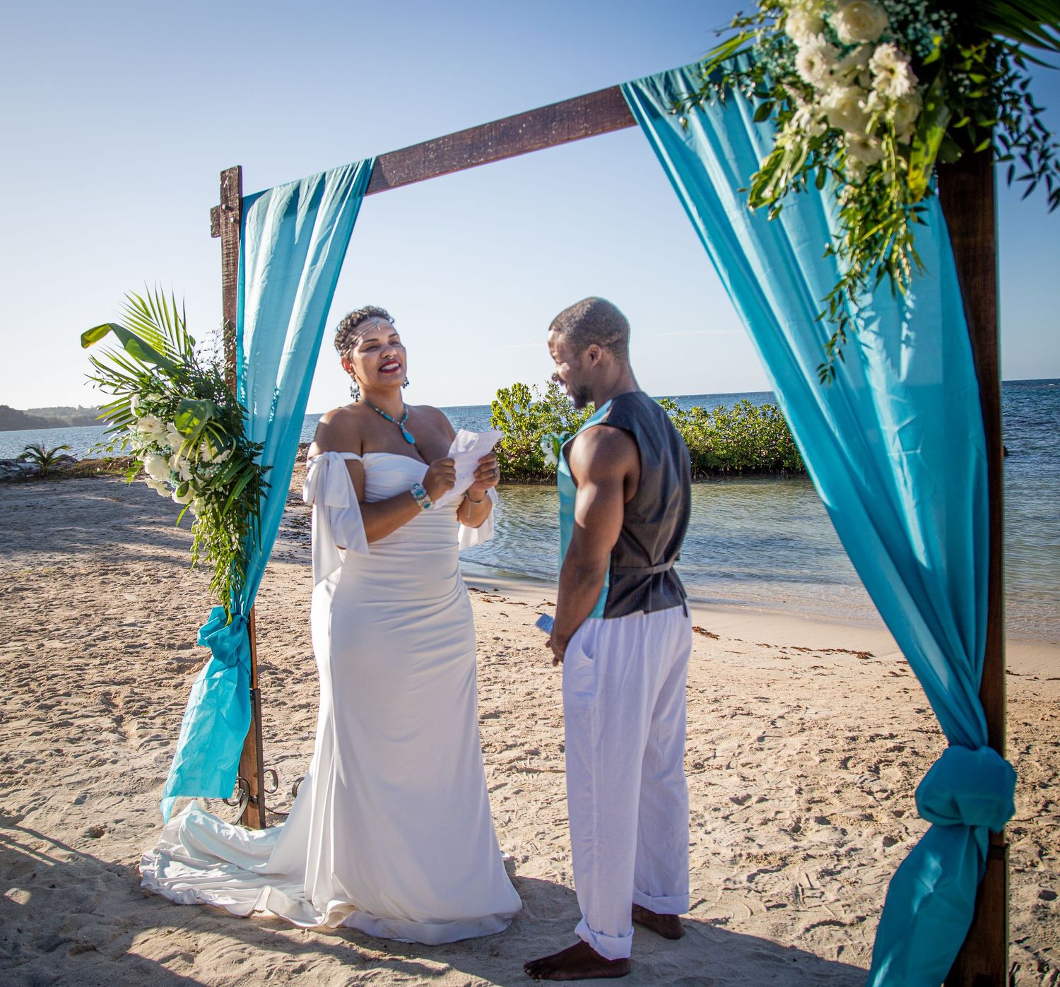 Beach wedding ceremony under wooden arch draped with turquoise fabric and floral decorations.