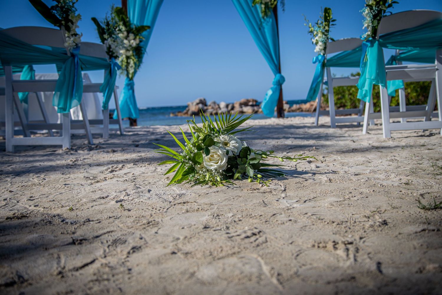 A beach wedding setup with turquoise fabric canopies and white chairs on sandy shoreline with ocean view.