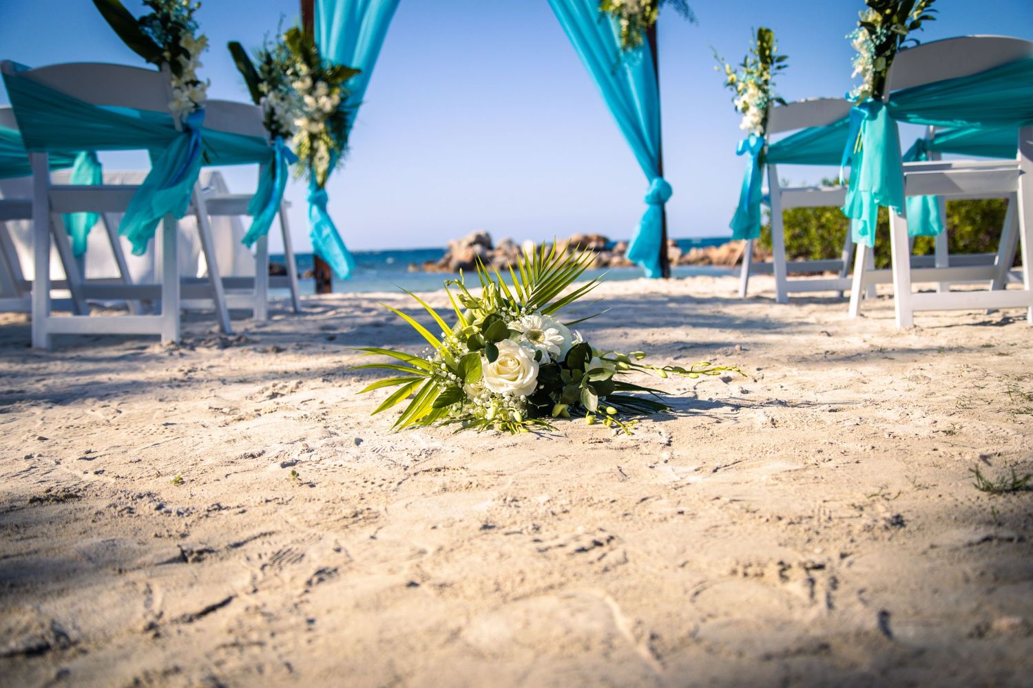 Beach wedding decor with turquoise fabric arch and white chairs on sandy shore.
