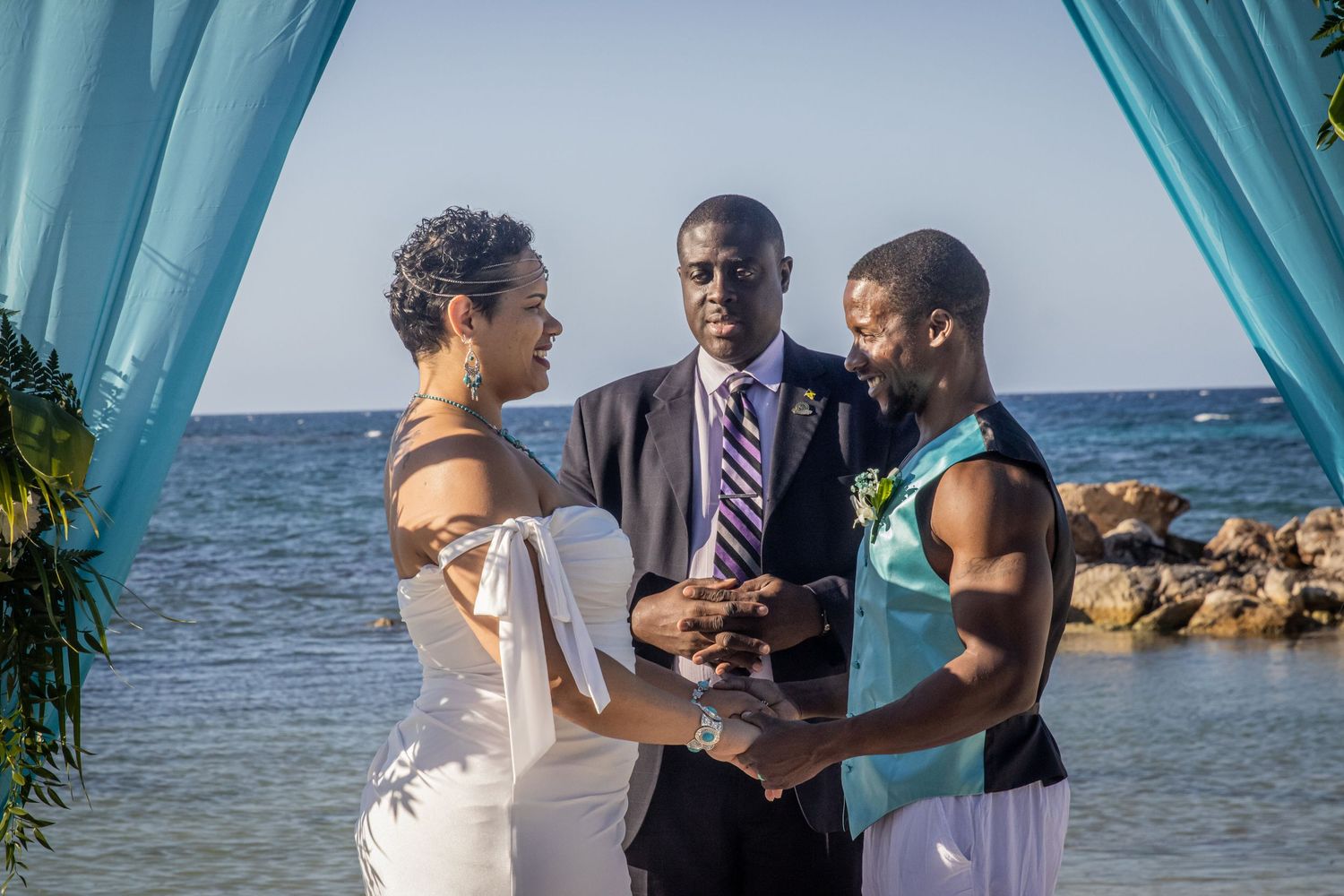 Wedding ceremony in progress under a turquoise beach canopy with ocean and rocky shoreline in background.