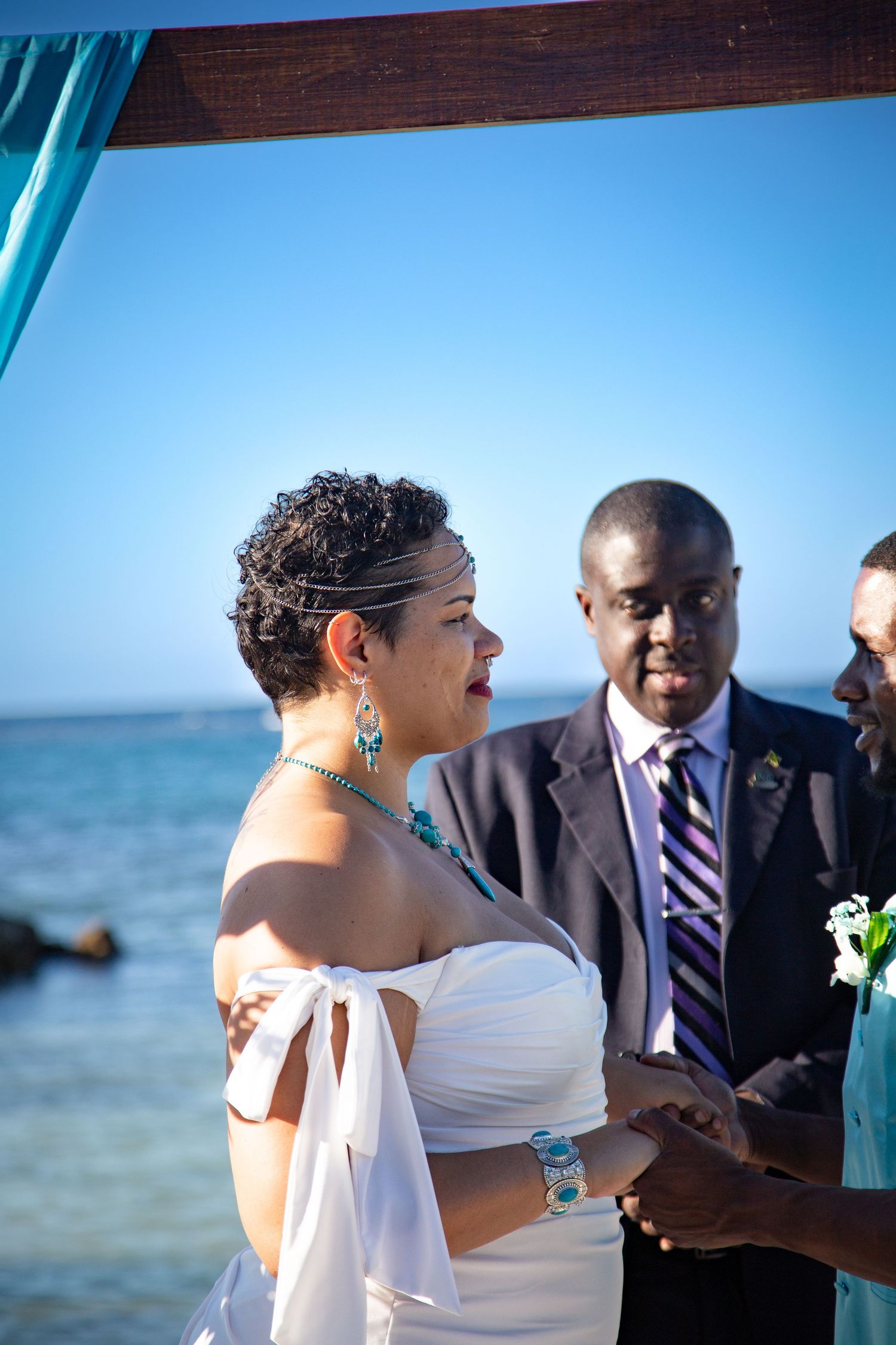 Beachside wedding ceremony with participants gathered under turquoise draped arch against blue ocean backdrop.