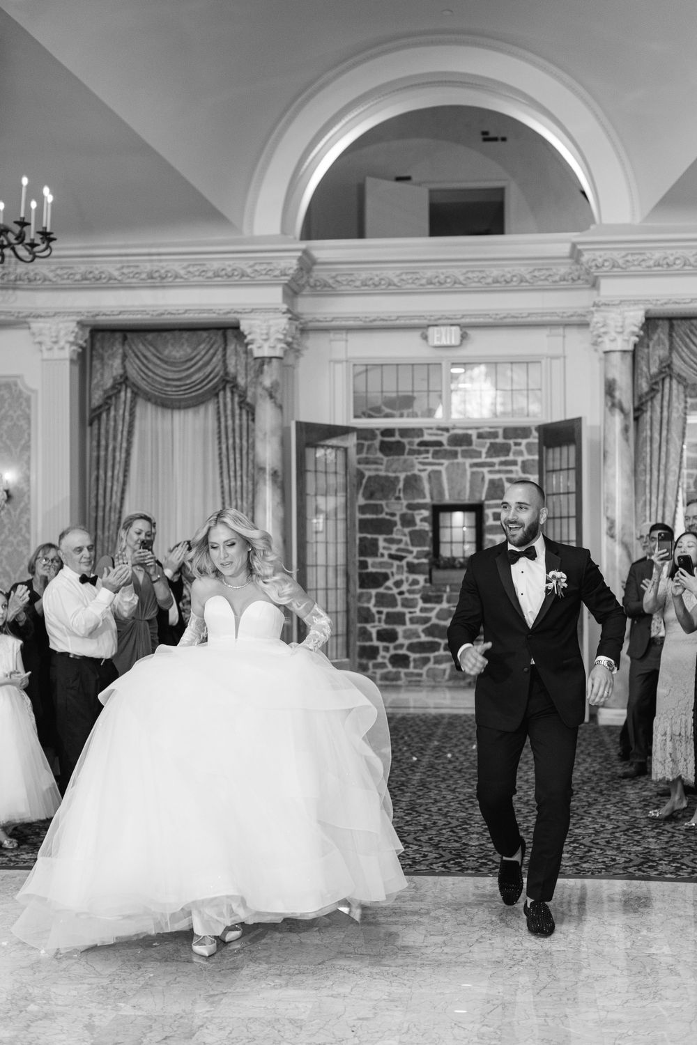 Couple makes grand entrance in ornate ballroom as guests look on during wedding celebration.