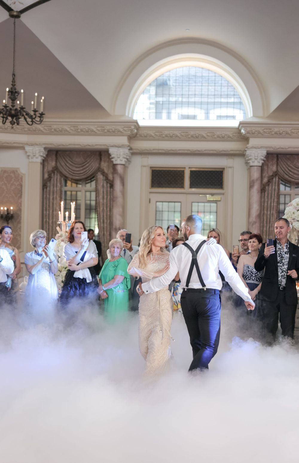 Guests dancing at an elegant wedding reception with fog effects and grand arched windows.