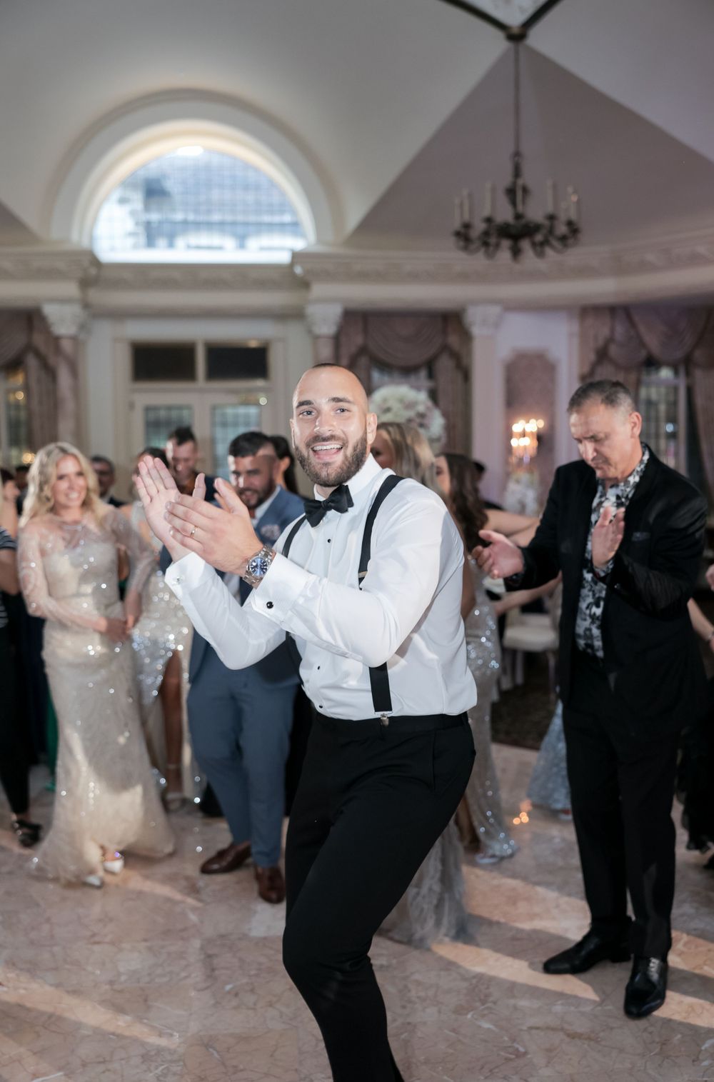 Festive wedding reception with guests dancing in an ornate ballroom with chandelier.