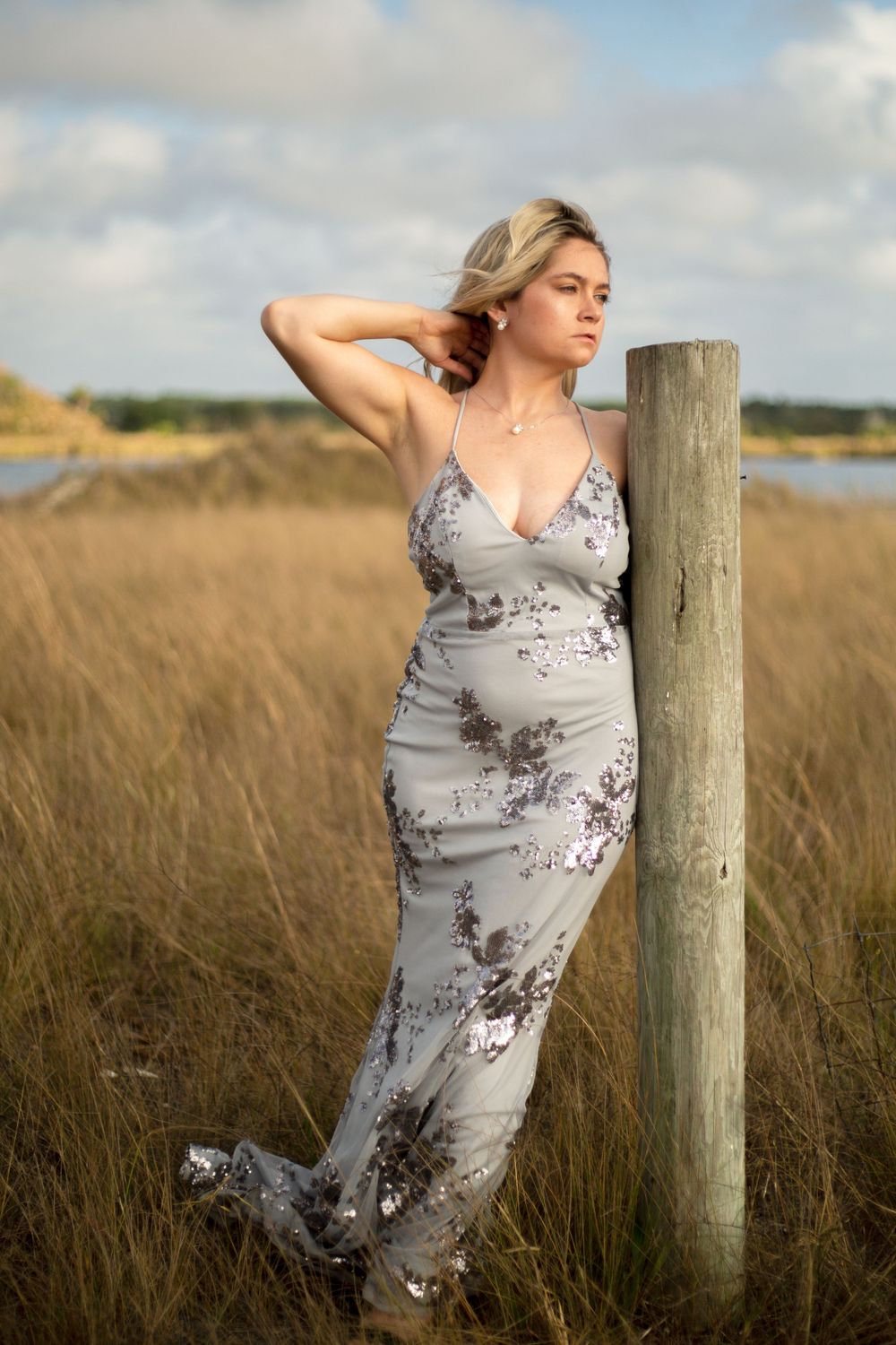 A woman in a floral dress poses against a wooden post in a golden coastal field.