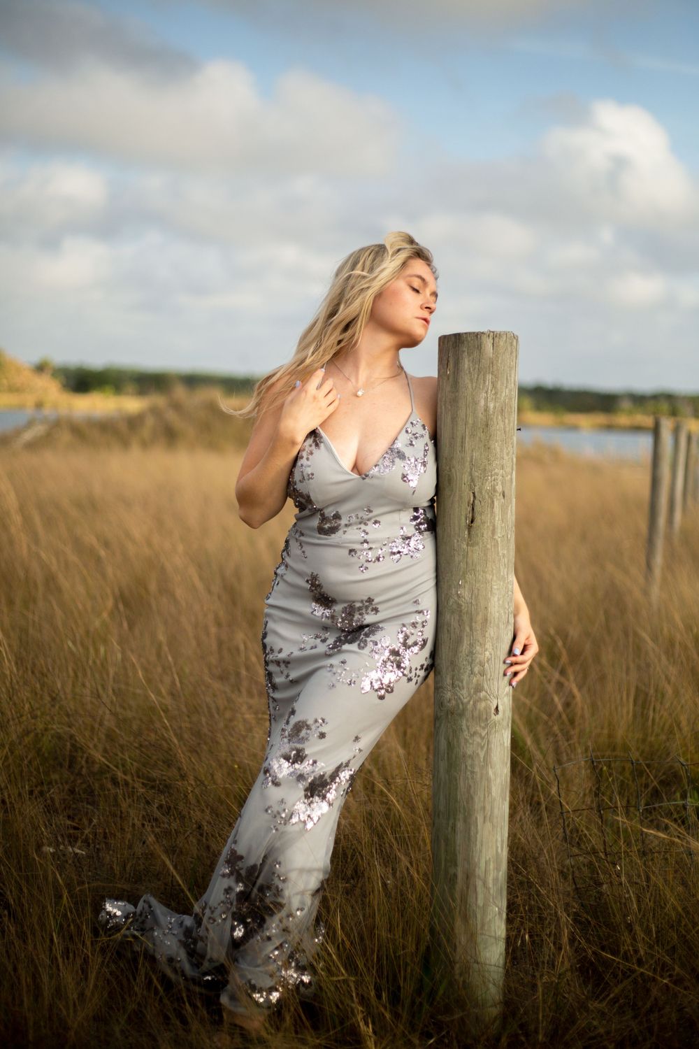 A woman in a flowing gray dress poses elegantly against a wooden post in a coastal field.