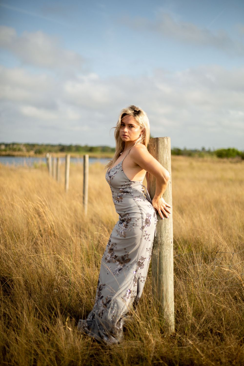 Woman in floral dress poses against wooden post in sunset-lit field of tall grass.