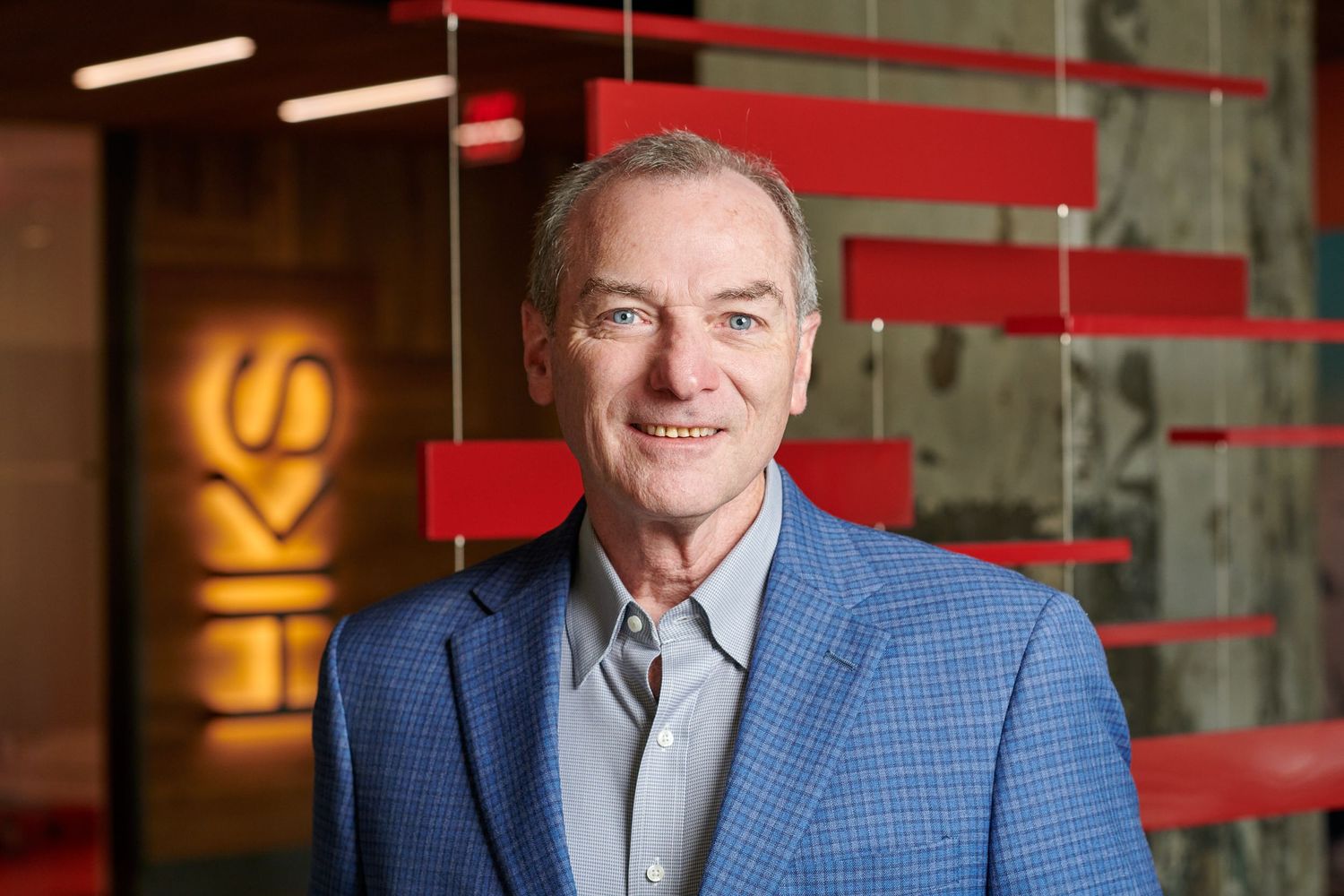 Professional portrait of person wearing blue checkered blazer in corporate setting with red accents.