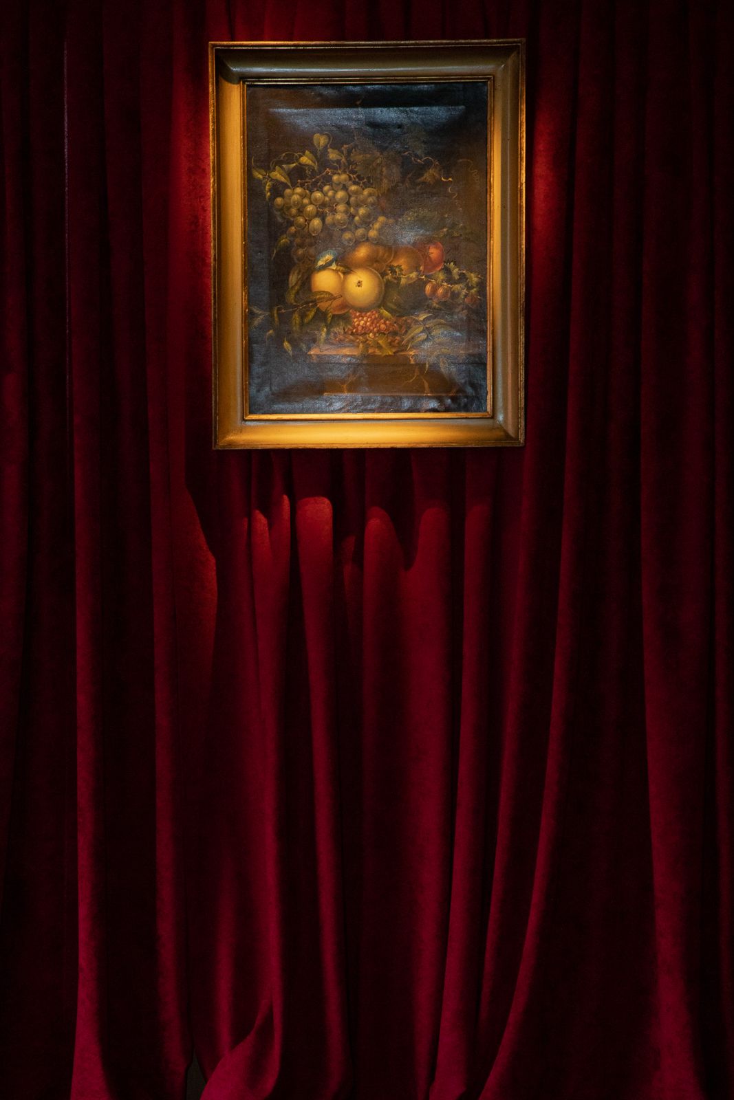 A gold-framed still life painting of fruit and flowers hangs on a wall with rich red velvet curtains.