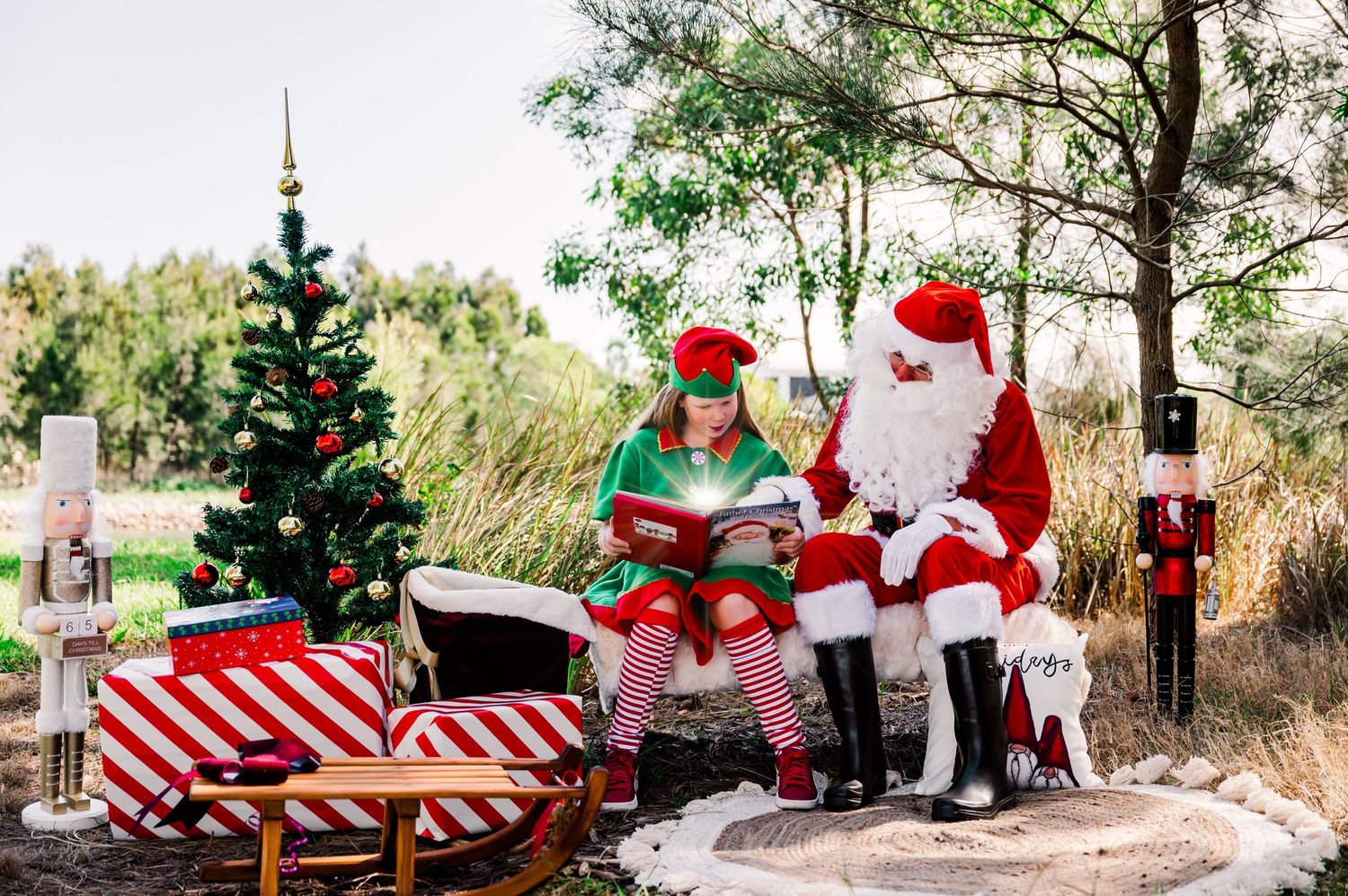 Santa Claus and helper read stories by decorated Christmas tree with wrapped presents outdoors.