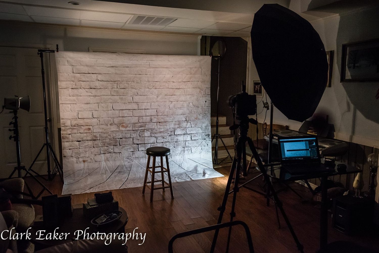 Photography studio setup with white brick backdrop, lighting equipment and wooden stool.