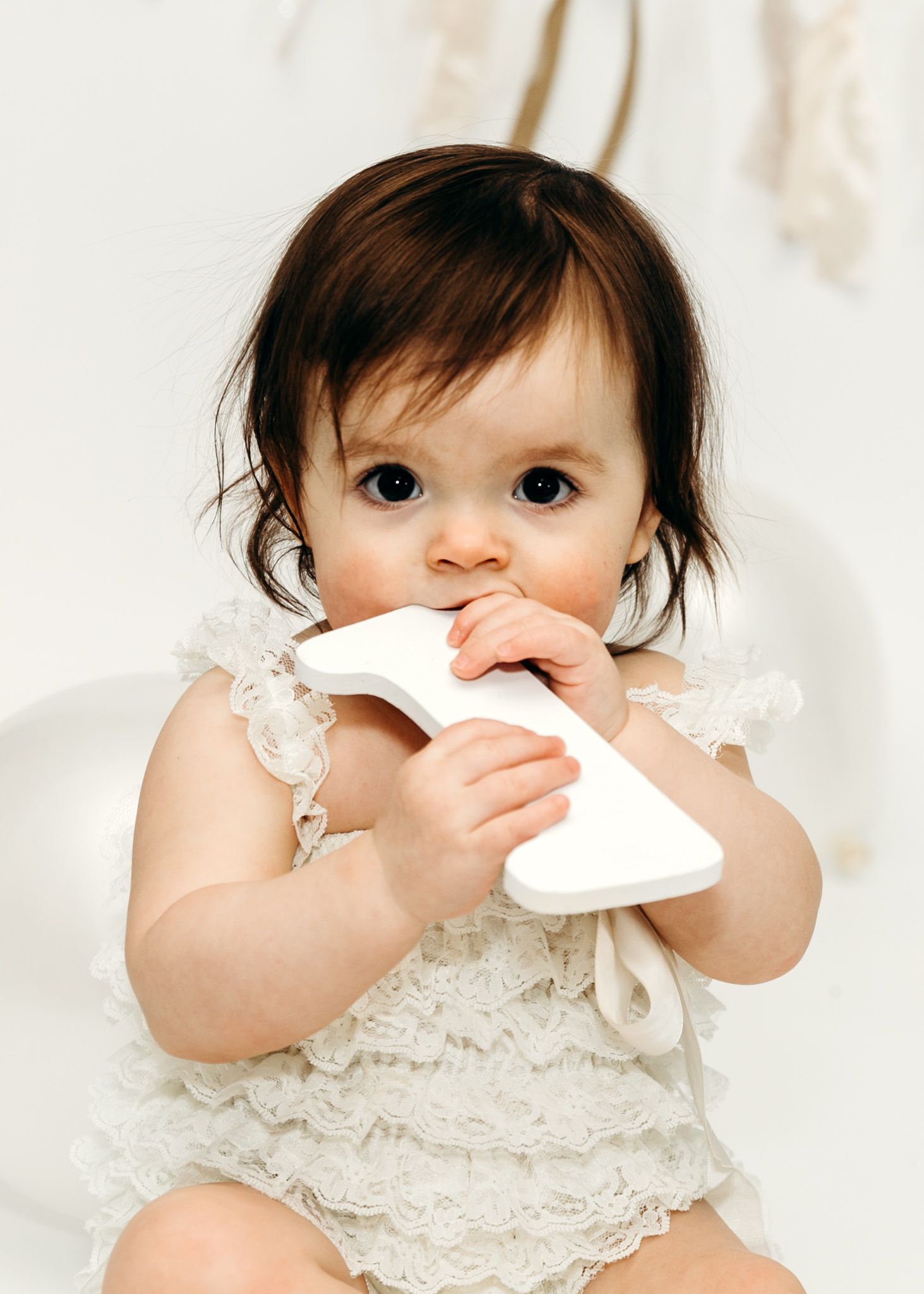 Adorable dark-haired baby in white lace romper chewing on number one cake topper.