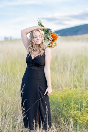 A black dress in a golden field holding wildflowers against mountain backdrop during sunset.
