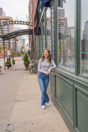 Woman in casual attire leans against green storefront window on city sidewalk.