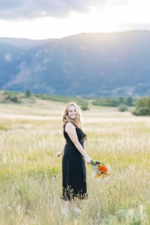 Standing in sunlit meadow wearing black dress and holding colorful bouquet.