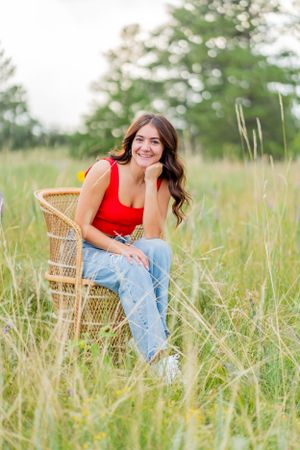 Woman in red top sits on wicker chair in tall grass field.