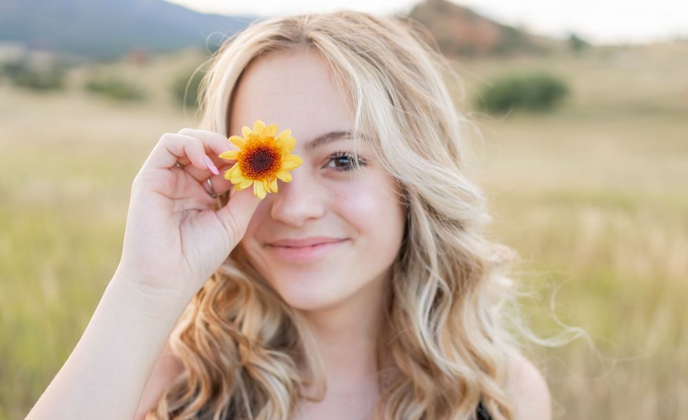 Playful portrait with a sunflower held up against mountain landscape.