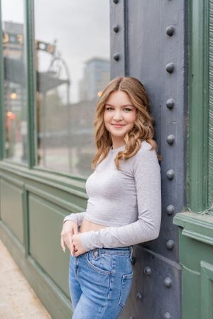 Woman in gray long sleeve top poses against industrial green and metal building exterior.