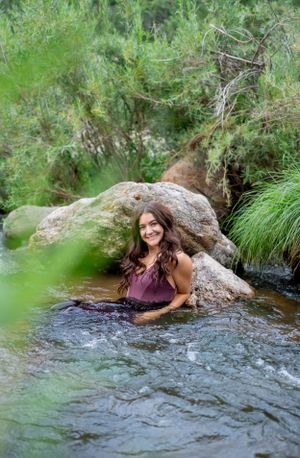 Figure lounging in a mountain stream surrounded by rocks and lush greenery wearing a purple dress.