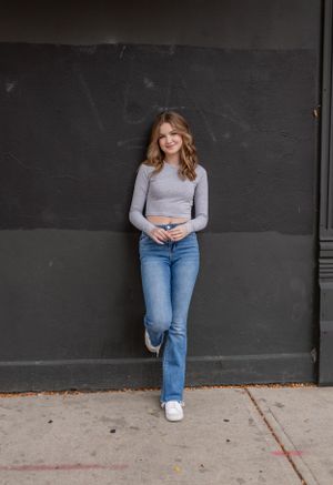 Woman in gray top and jeans poses against dark industrial wall in urban setting.