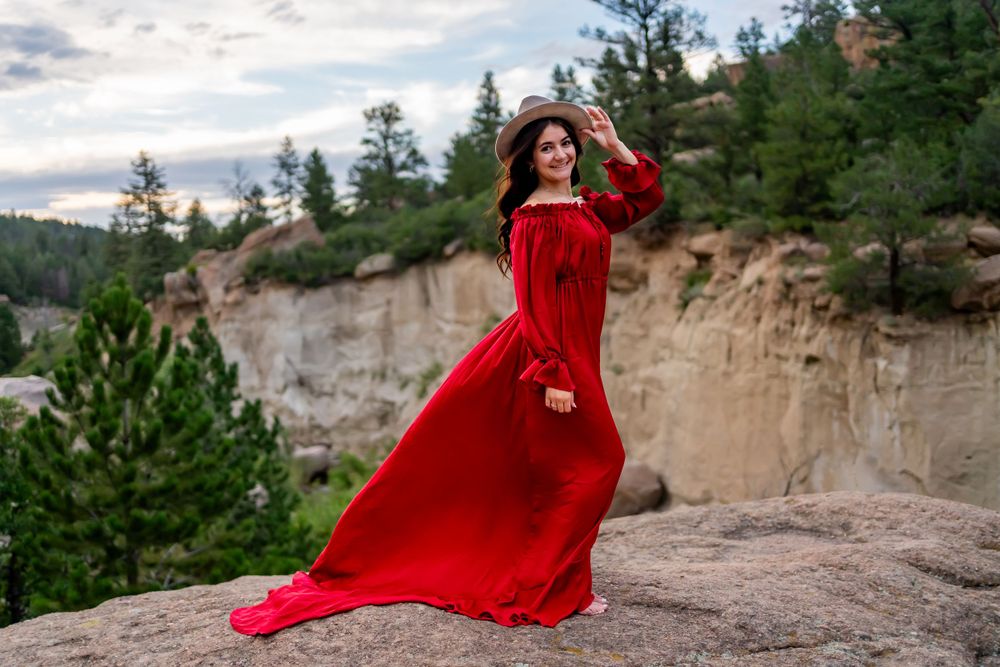 Woman in flowing red dress stands on cliff edge with mountain backdrop.
