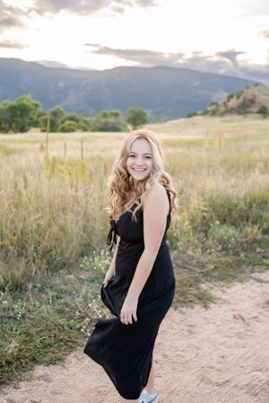 Woman in black dress smiles while standing on dirt path with mountains in background.