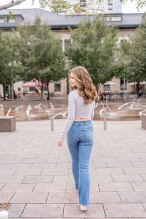 Woman in light gray crop top and flared jeans poses outdoors in an urban plaza setting.