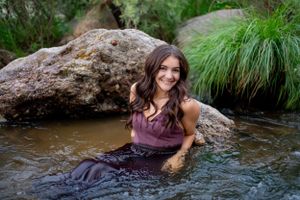 Person sitting in crystal clear stream water against large boulders wearing a flowing purple dress.