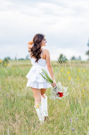 White dress and boots in a summer field holding wildflowers and walking through tall grass.
