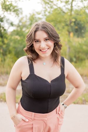 A person in a black top and pink pants poses outdoors with a confident smile.