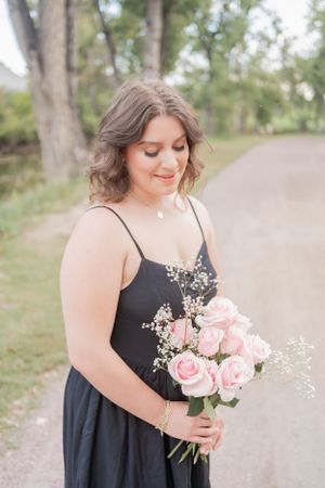 A person holds a bouquet of pink roses and baby's breath while wearing an elegant black dress outdoors.