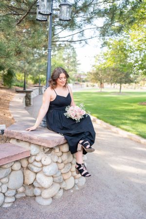 A summer garden portrait with a black dress and bouquet while sitting on a stone wall.