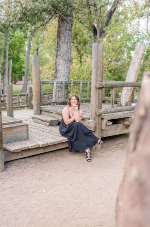 A person in a long black dress sits elegantly on a wooden deck pathway surrounded by trees in a park setting.