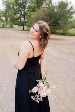 A person in a black dress holds a bouquet of pink roses on a path surrounded by greenery.