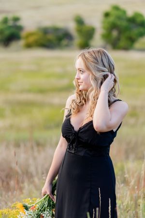 Figure in black dress stands among wildflowers in mountain field at golden hour.