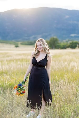 Walking through golden field in black dress with bouquet at sunset.
