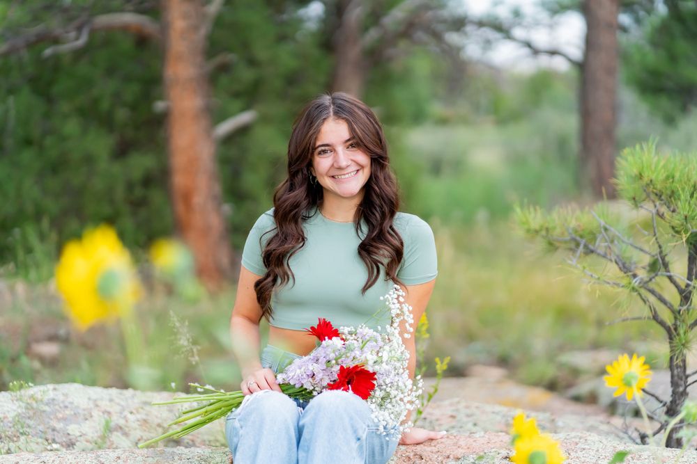Woman in mint green top holds flowers while sitting on rocks outdoors.