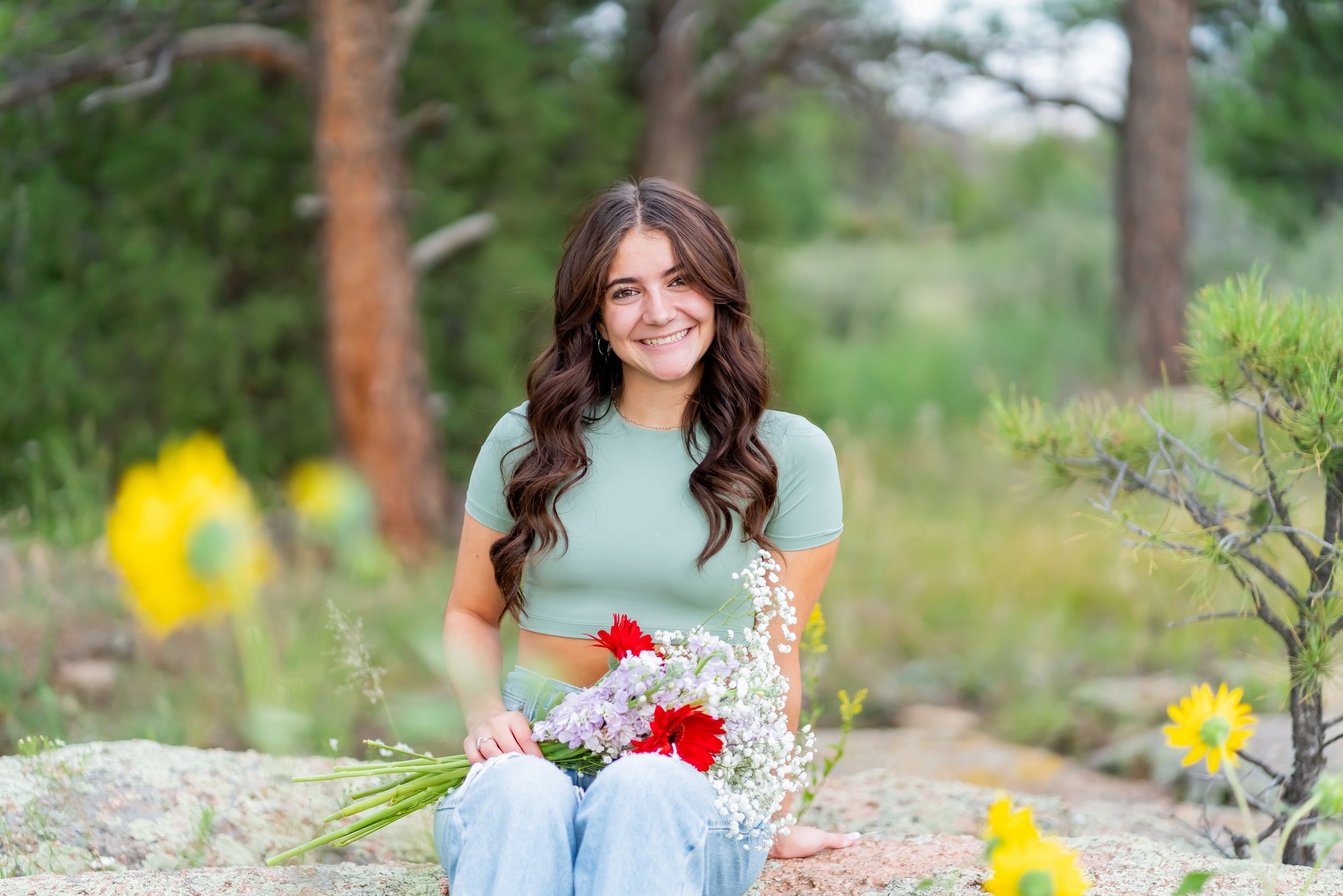 Woman in mint green top holds flowers while sitting on rocks outdoors.