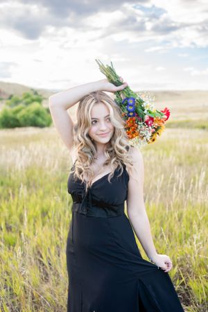 Person in black dress poses with colorful wildflower bouquet in mountain meadow at sunset.
